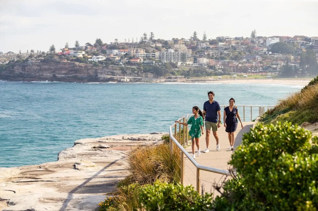 Family walking the boardwalk between Bondi to Bronte Beach with beach and cliff-side housing in the background