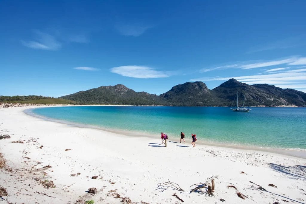 Hikers walking along the white-sand beach of Wineglass Bay with a sailing vessel in the background