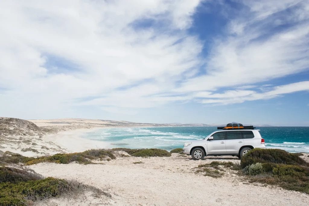 A 4WD with surfing gear parked on a ledge overlooking a white-sand beach