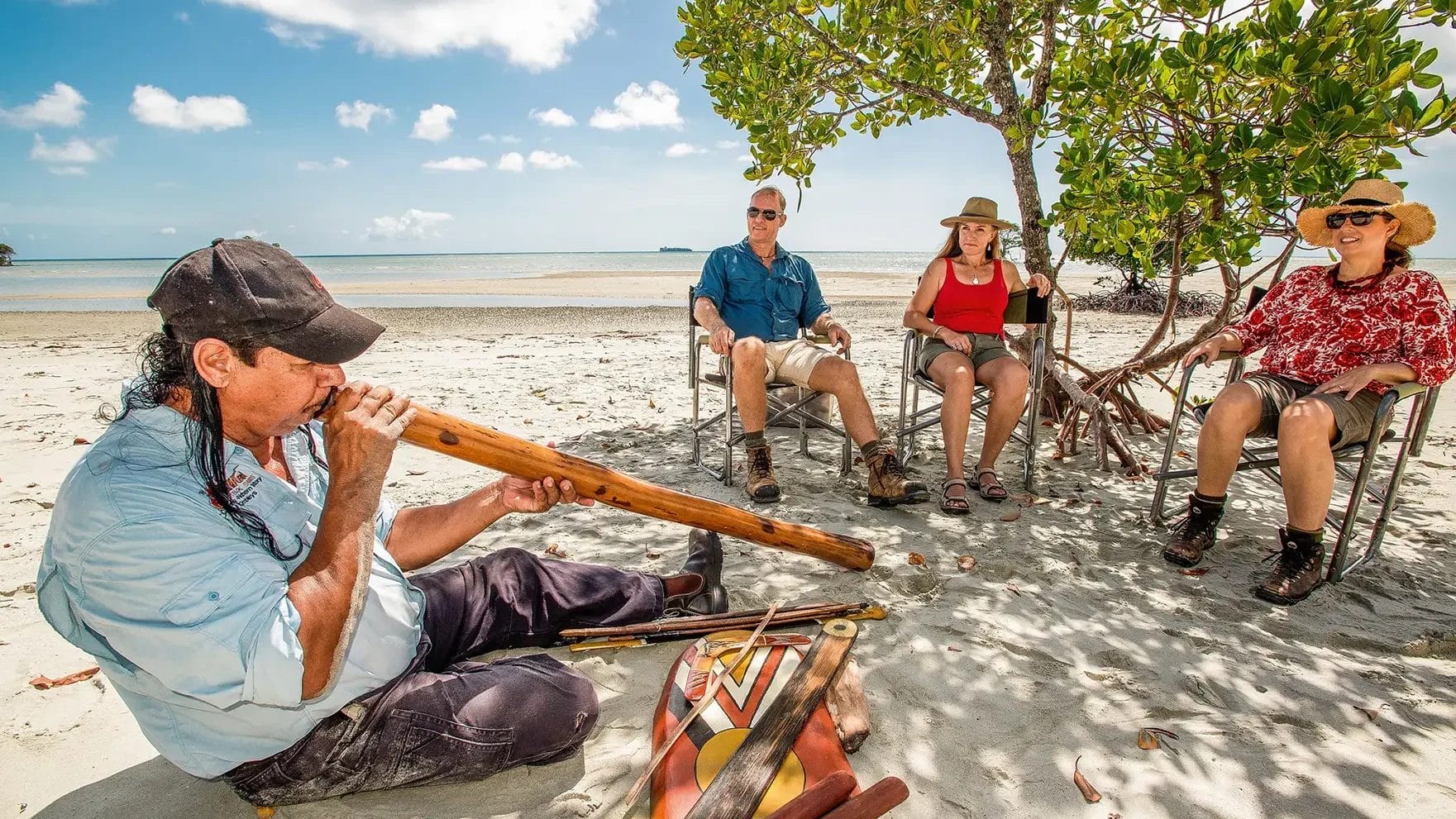 Three travellers watching a didgeridoo performance on a white sand beach